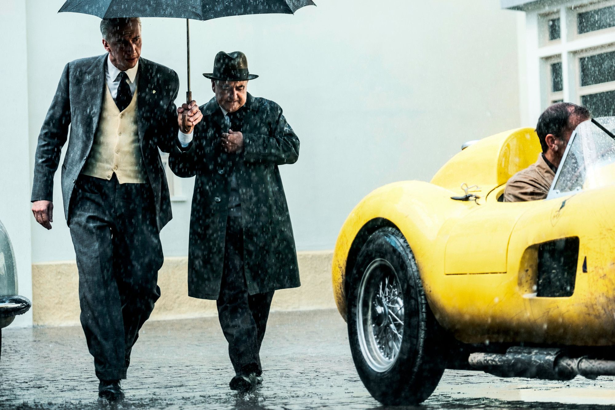 Adam Driver as Enzo Ferrari in Ferrari walking under an umbrella in the rain with another man next to a yellow 1950s race car