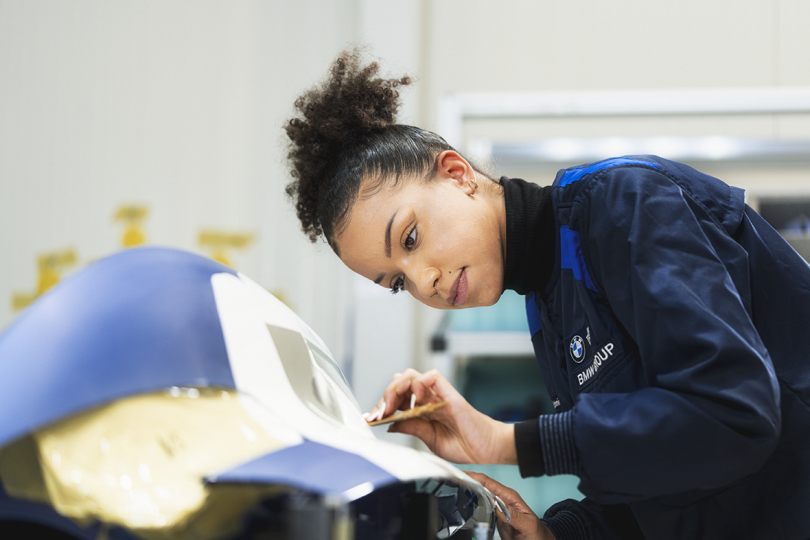 brown-skinned woman leaning over painting piece of car exterior