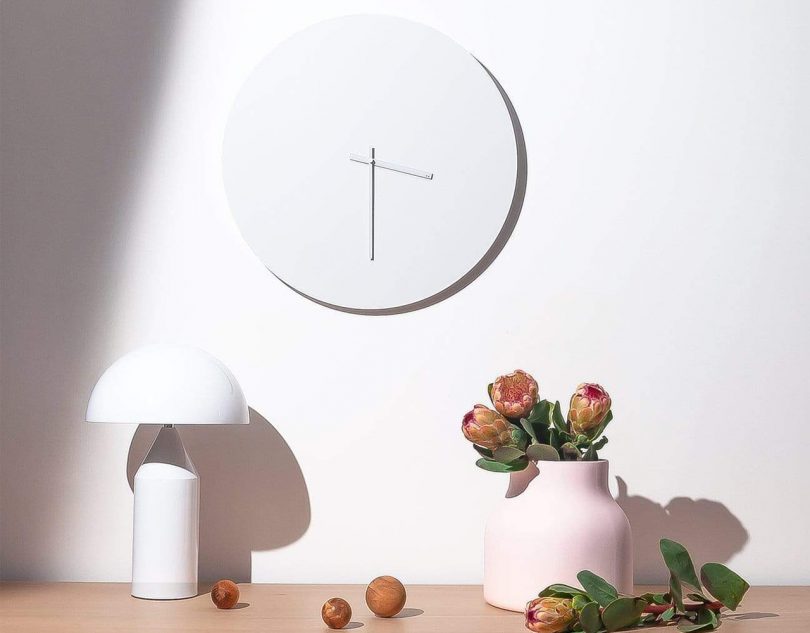 TOO designs minimal wall clock in white hanging over a table with a white lamp and vase full of flowers