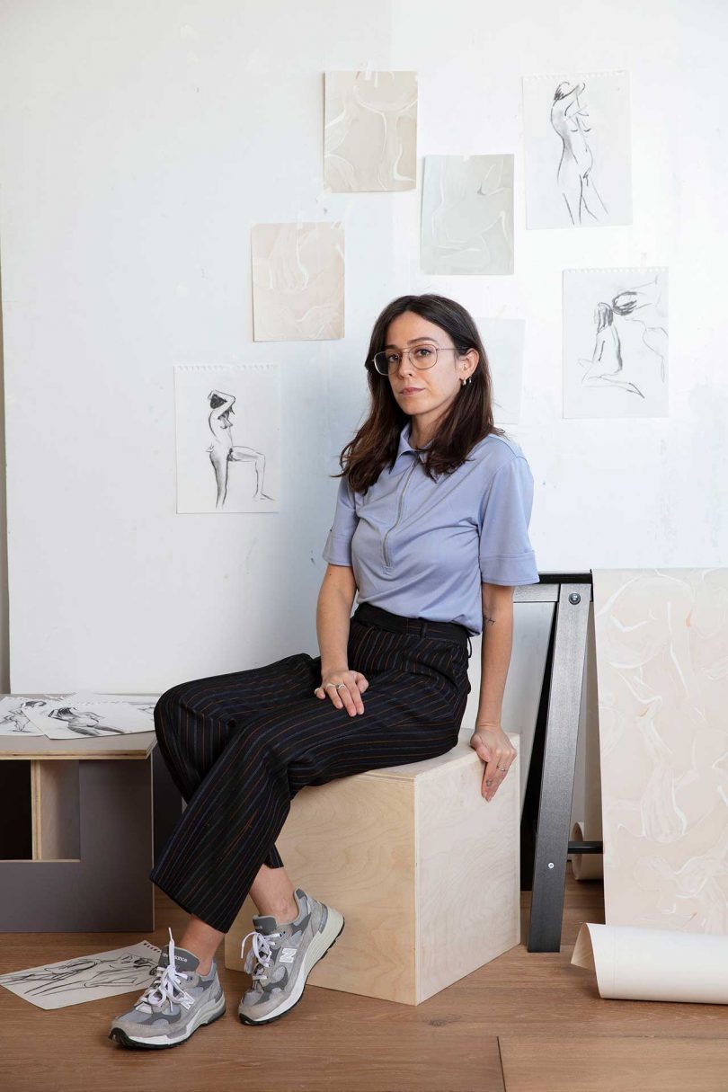 Person sitting on wood cube with sketches on wall behind them
