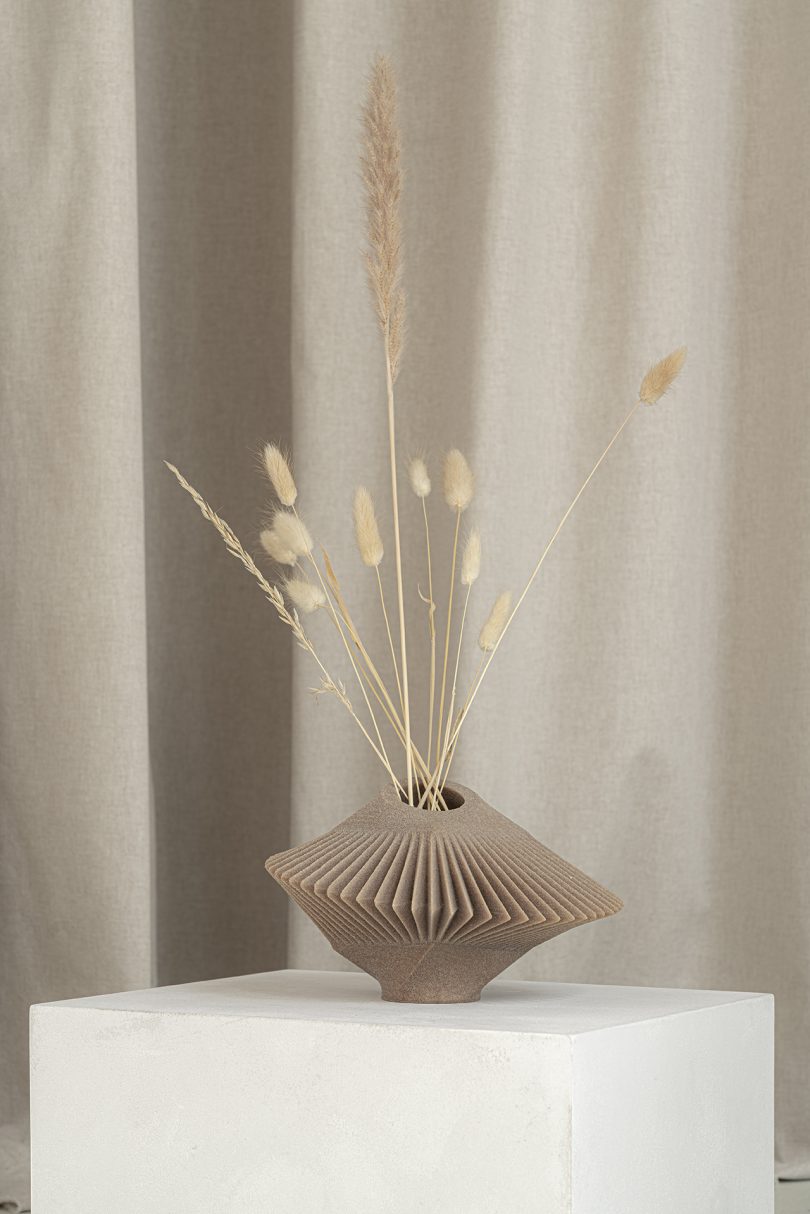 light brown abstract vessel with dried stems sitting on white pedestal in front of light fabric