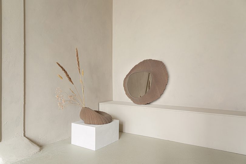 light brown abstract vessel sitting on white pedestal and light brown mirror sitting on a ledge leaning against light wall