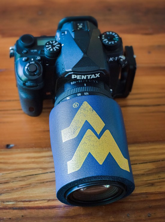 A West Virginia Mountaineers beer cozy around a lens to hold hand warmers in place. For many lens, you might need to cut this and either apply Velcro strips to close it back up, tape it with gaffer's tape, or tie rubber bands around it.